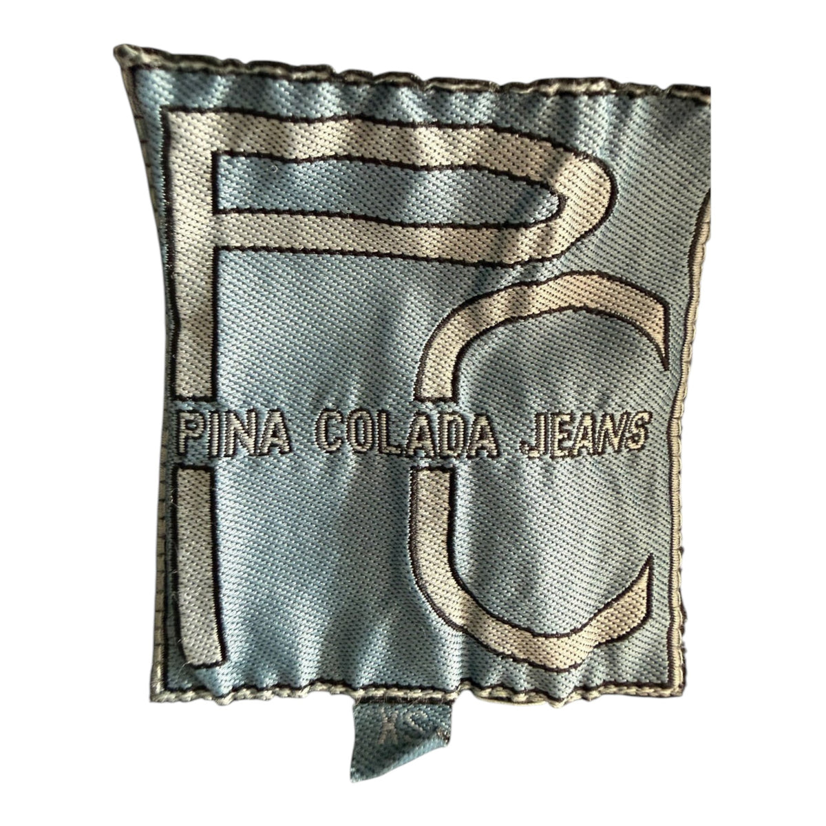 Metka odzieżowa marki Pina Colada Jeans z logo PC na jasnoniebieskim tle w sklepie TrickandChic