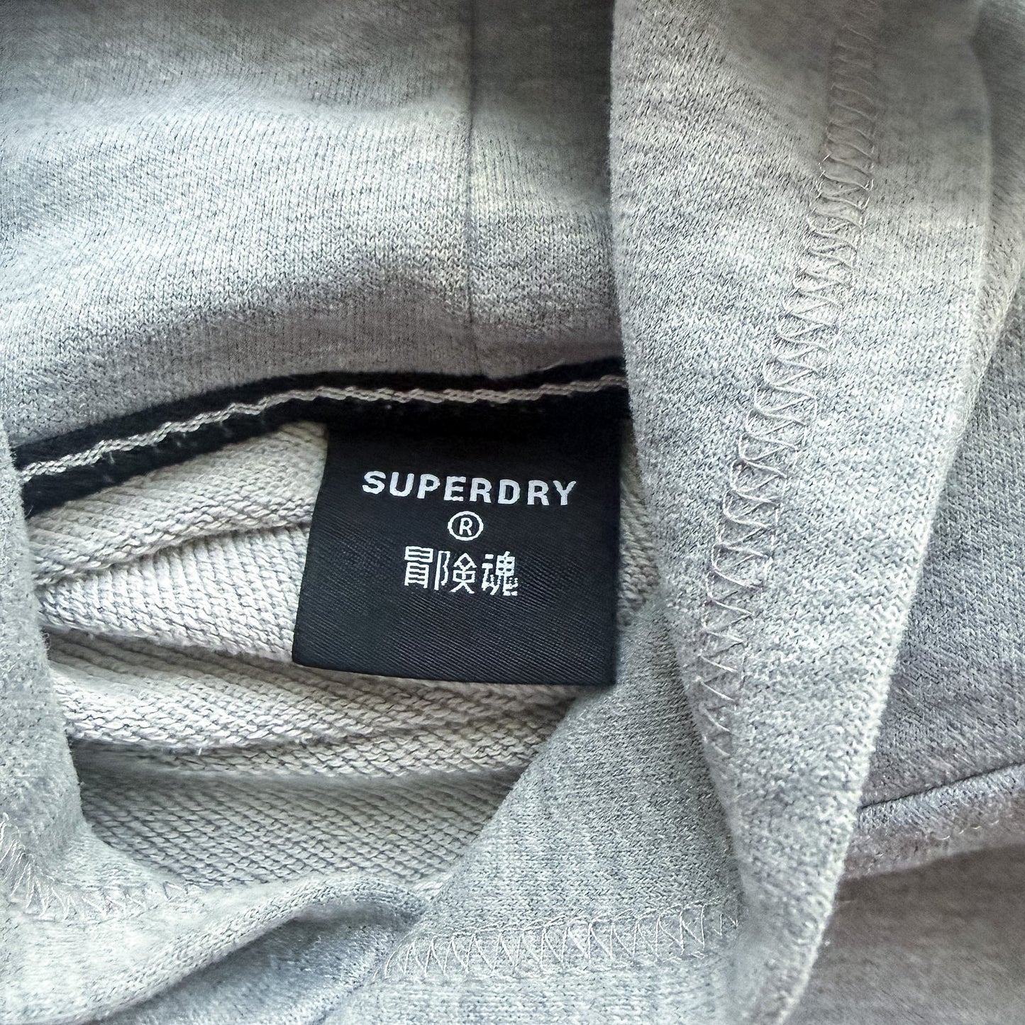 Metka marki Superdry wewnątrz szarej bluzy z kapturem TrickandChic z widocznym logo i japońskimi znakami